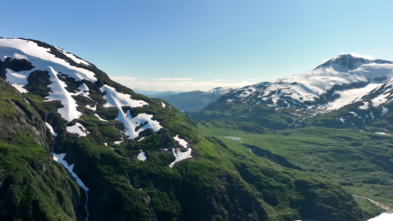 Lush Green Slopes and Snow Patches of Alaskan Mountains. A bright summer scene highlights the dramatic contrast between the steep, lush green, forested slopes of Alaskan mountains