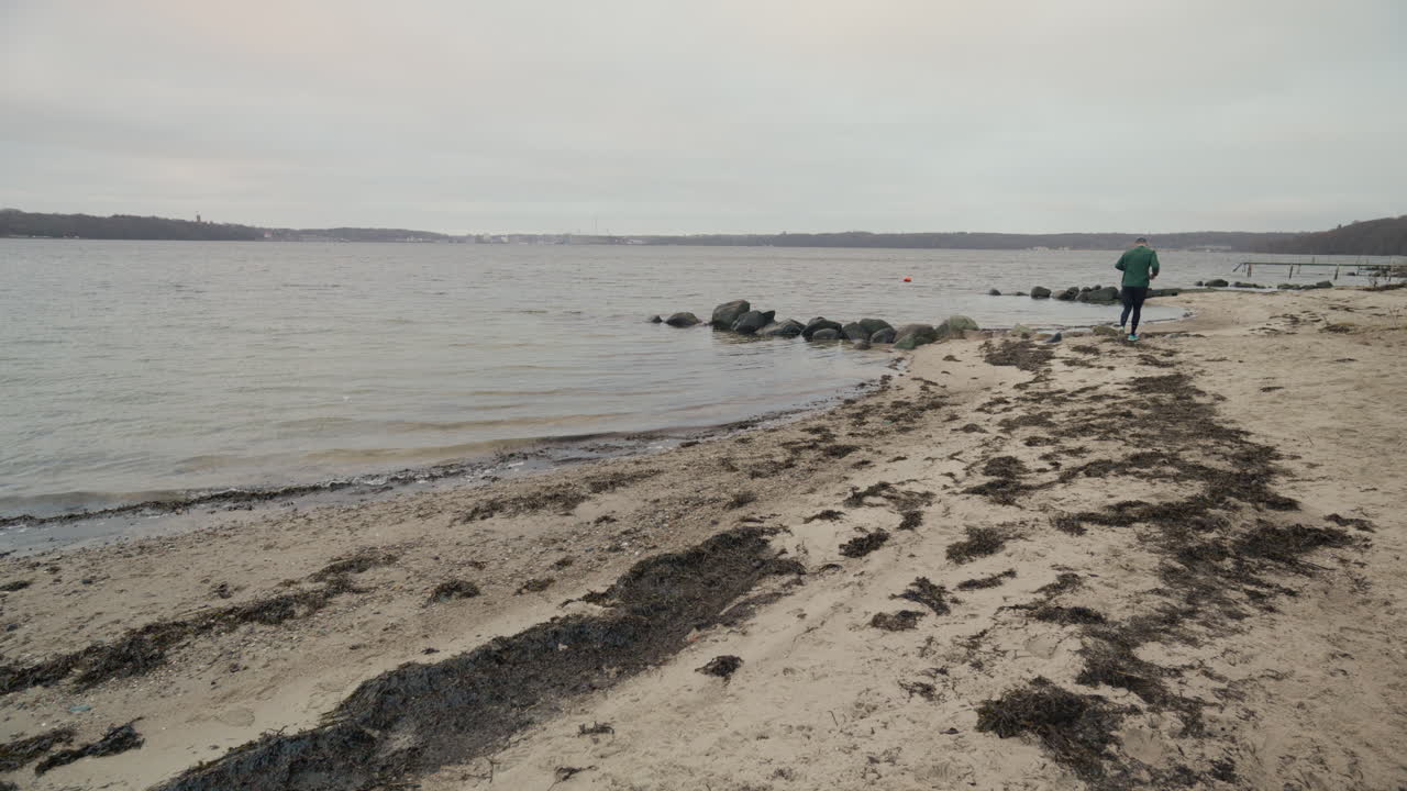 Jogger som løper bort fra kameraet på en vinterstrand nær vannet en overskyet dag.