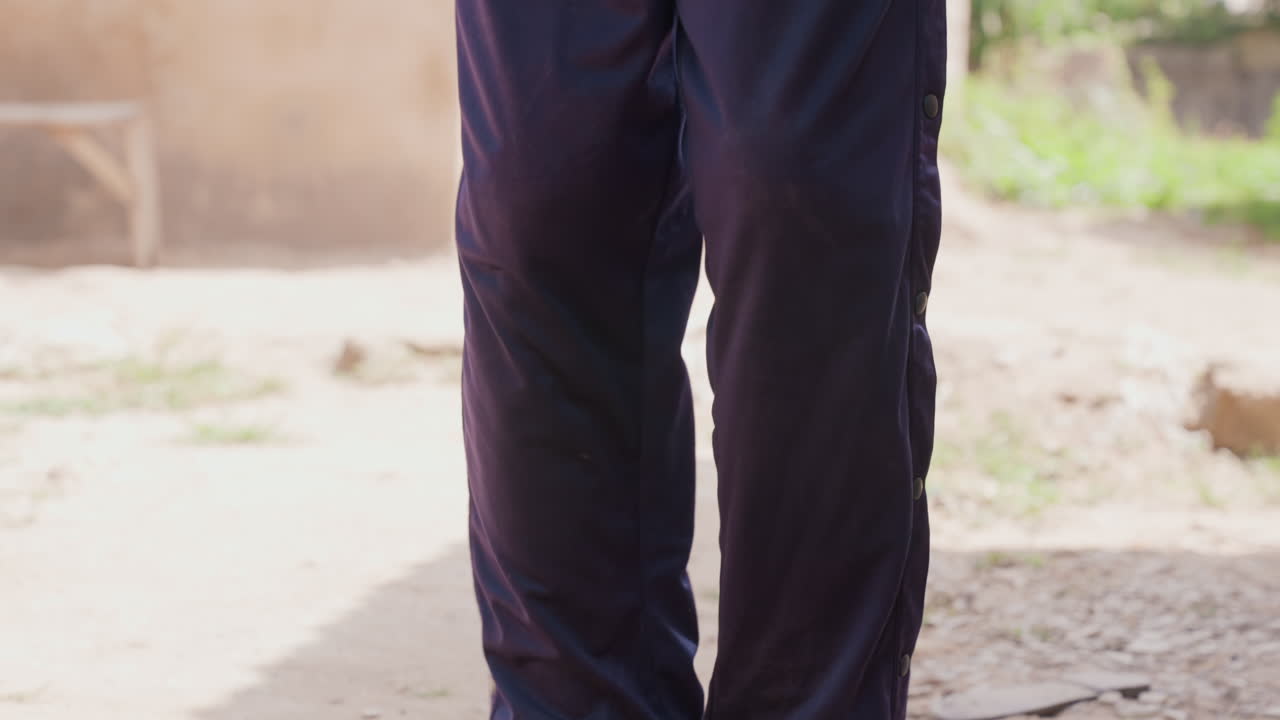 Black athlete dribbling ball on dirt, closeup legs in dark tracksuit, worn red basketball bouncing with controlled footwork, slowmotion rhythm, gritty urban court, warm sunlight and shadow, solitary