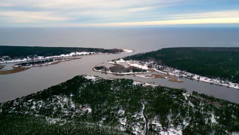 Vista-Aérea-Invernal-De-Un-Río-Sinuoso-Rodeado-De-Un-Exuberante-Bosque-Verde,-Con-El-Estuario-De-Lielupe-En-El-Mar,-El-Mar-Báltico-En-Colores-Pastel-En-La-Distancia