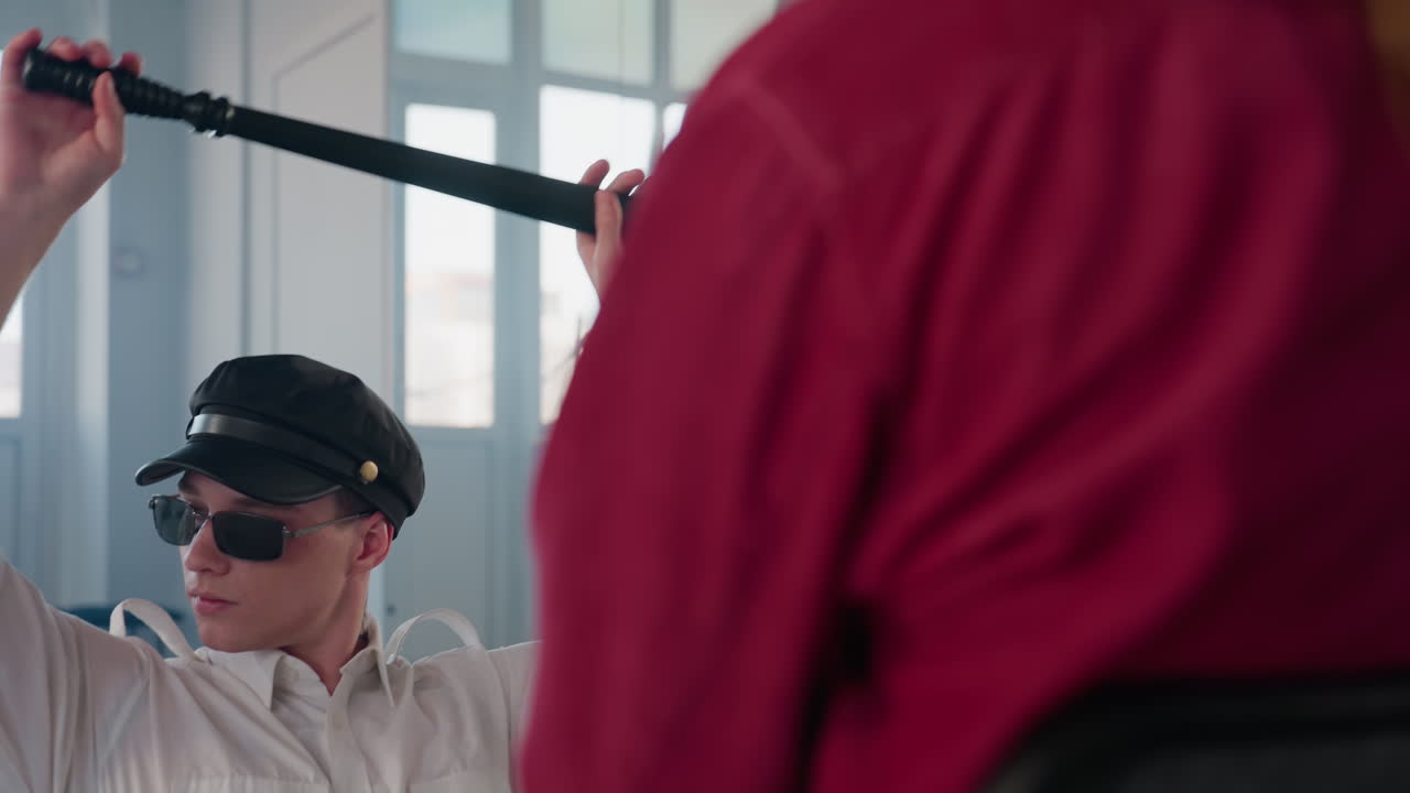rear view of seated woman in red shirt facing male stripper wearing cap sunglasses sliding baton behind head while dancing squatting near mirror in bright minimalist studio with window reflections