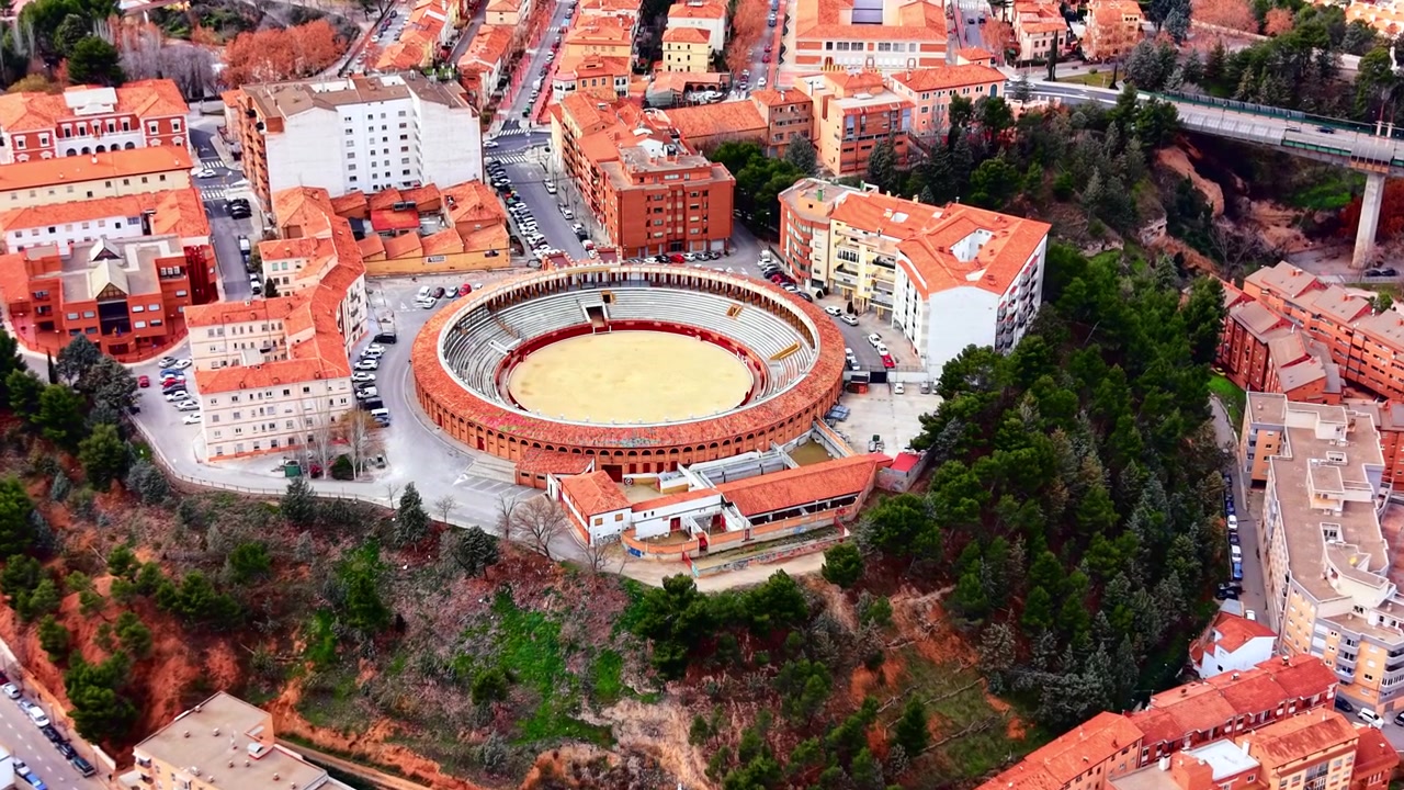Vista dall'alto di un'arena per la corrida circondata da edifici residenziali, Teruel. Arena circolare di sabbia con porticato ad archi, tetto in tegole rosse, collina di pini e un burrone con pilastro di viadotto ai margini della città.