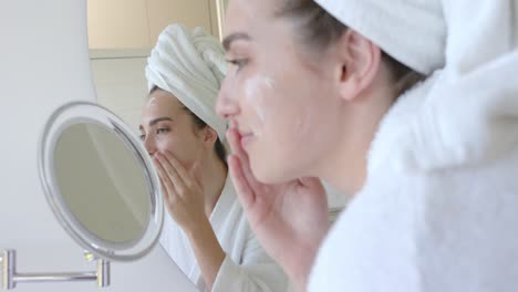 Caucasian-woman-with-towel-on-head-applying-cream-on-face-in-bathroom-in-slow-motion