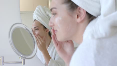 mujer caucásica con una toalla en la cabeza aplicando crema en la cara en el baño en cámara lenta