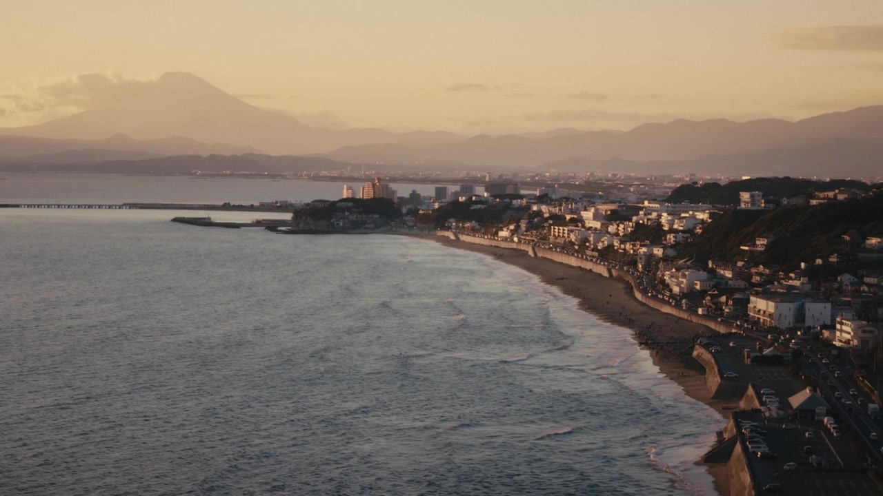 Veduta aerea della costa curva di Shichirigahama al tramonto, Kamakura, Giappone, con la luce dorata che si riflette sulle onde e le case in collina che costeggiano la riva.
