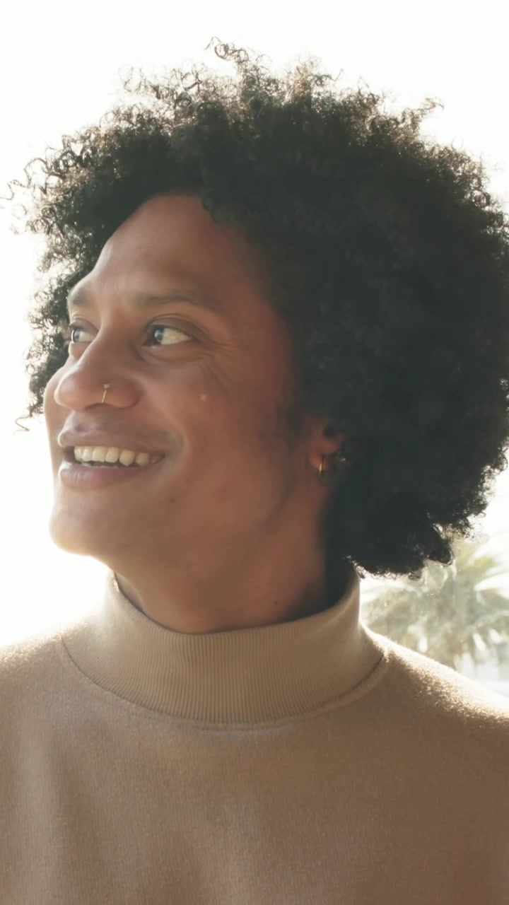 Vertical video: Smiling man turning left looking up at window in tan sweater nose ring palm trees