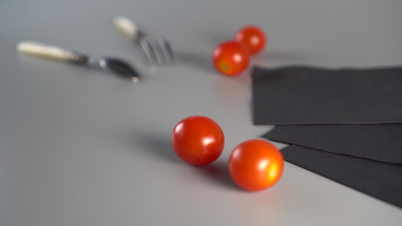 Red ripe cherry tomatoes fall from above on a gray background, still life with a spoon, a fork and three black napkins.