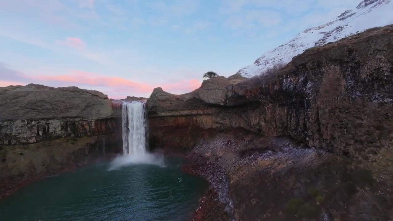 Stunning aerial footage of the Salto del Agrio waterfall plunging into a pool. Features a pink sunset sky and snowy mountains in Neuquén, Argentina.
