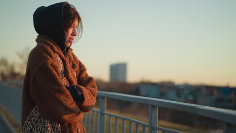 a close-up of a tearful girl wearing a brown coat and hoodie, standing near a metal railing. her hands are folded in her arms as she gazes thoughtfully into the distance, reflecting a sense of sadness