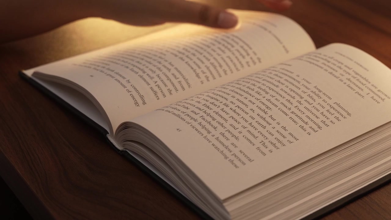 A woman's hand turns pages of an open book resting on a dark wooden table, bathed in warm golden lamp light, creating a cozy intimate reading atmosphere with soft bokeh in the background.