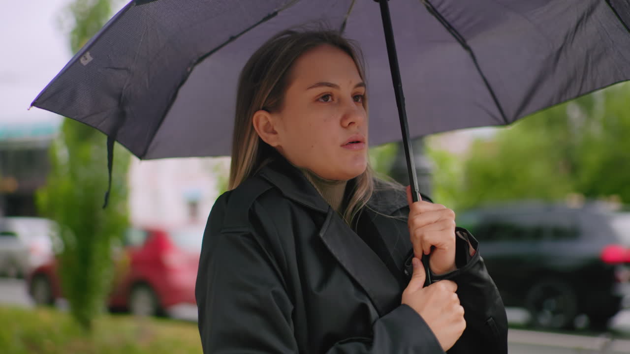 Gorgeous girl in black trench coat holding umbrella with both hands standing near bench on city sidewalk feeling so cold with blurred cars and buildings in background showing calm thoughtful outdoor lifestyle