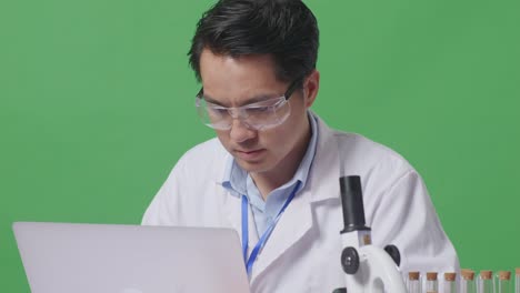close up of asian man scientist typing on a laptop while working on the table with microscope and test tube in the green screen background laboratory