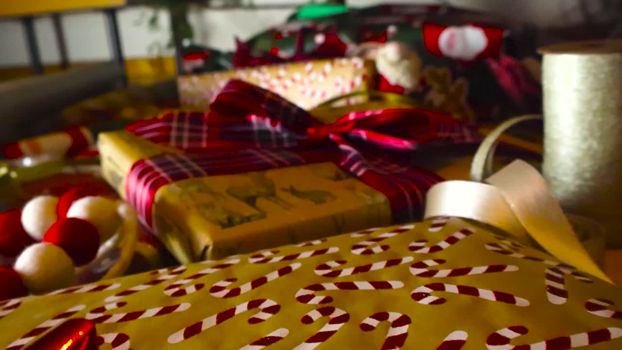 Close up footage gliding over candy cane and festive wrapped Christmas presents under holiday tree on the ground. Foreground is in sharp focus and background blurred. Red ribbons, wrapping paper rolls