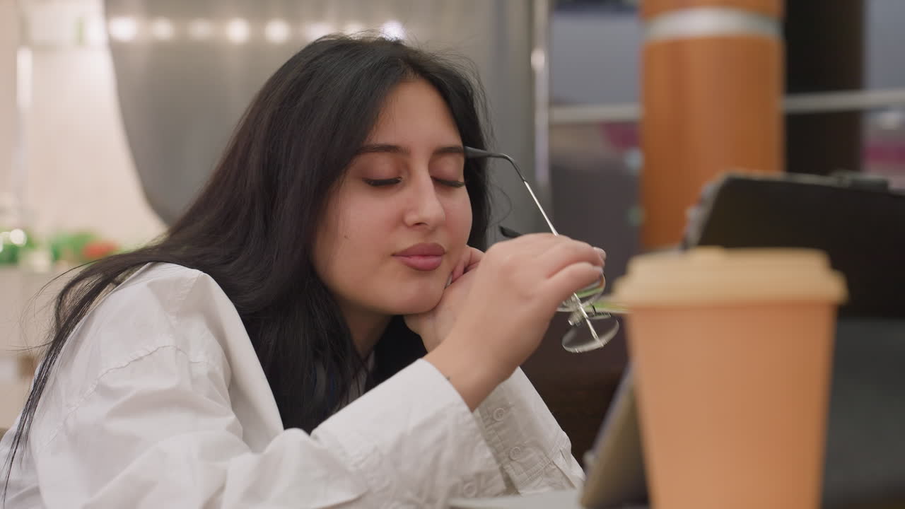 Elegant young woman seated at table gently removes glasses while staring at tablet with soft expression, indoor environment with warm lighting and takeaway coffee cup in foreground