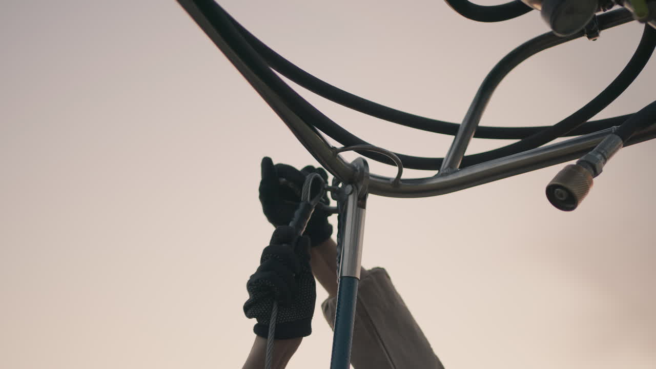 close up of gloved hands detaching metal hook from steel cable assembly against fading sky backdrop highlighting manual operation and tool release mechanism in industrial lifting equipment setup
