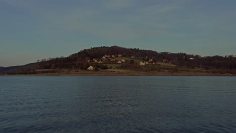 Flying-Above-Calm-Lake-Towards-Mountainous-Lake-Shore-Village-At-Dusk