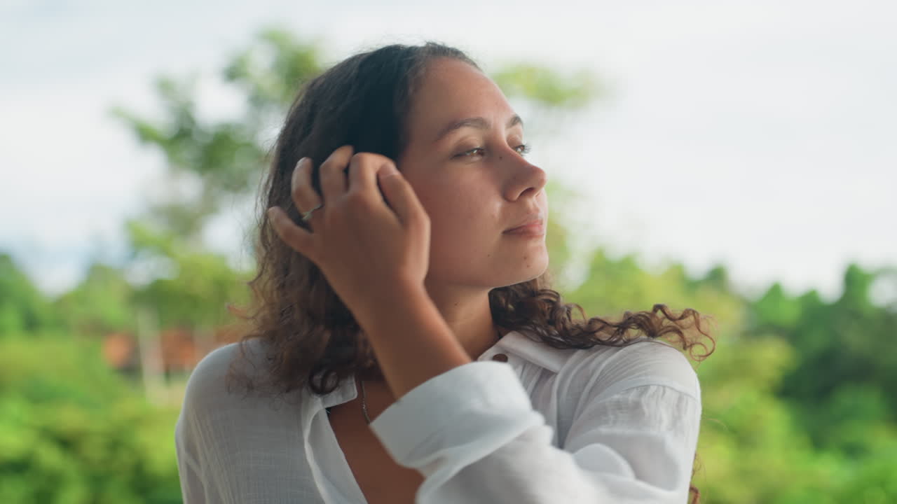 Retrato de primer plano con fondo de vegetación isleña, emoción sutil, dedos con cabello rizado tocando mechones, luz suave, detalle de joya, expresión contemplativa, toma íntima de belleza natural para estilo de vida.