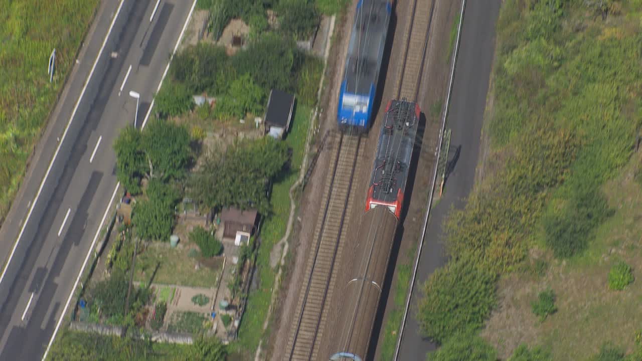 una toma aérea de un dron muestra un tren vibrante, adornado con vagones azules y amarillos, corriendo a lo largo de una vía paralela a una carretera bulliciosa, con un río sereno y casas pintorescas en el fondo