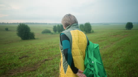 back view of woman wearing yellow and green jacket removing her green raincoat and preparing to pack it on grassy field under cloudy sky, with trees and vast landscape in the distance