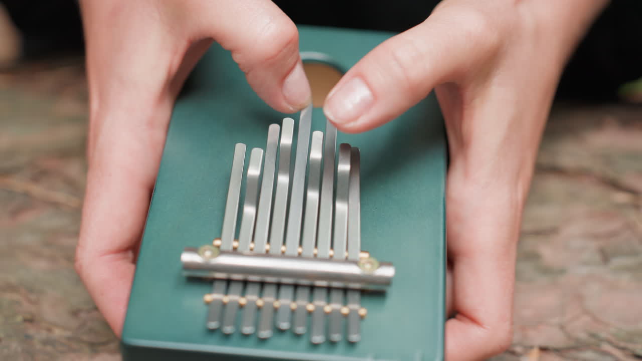 Extreme close up of hands in dark outfit delicately plucking metal tines on small green thumb piano, showing rhythm, texture, and emotion, evoking sense of calm creativity