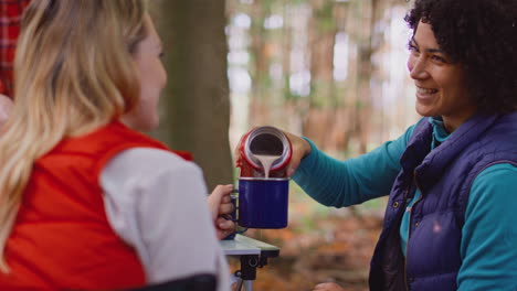 woman pouring hot drink from flask as female friends on camping holiday cook meal together