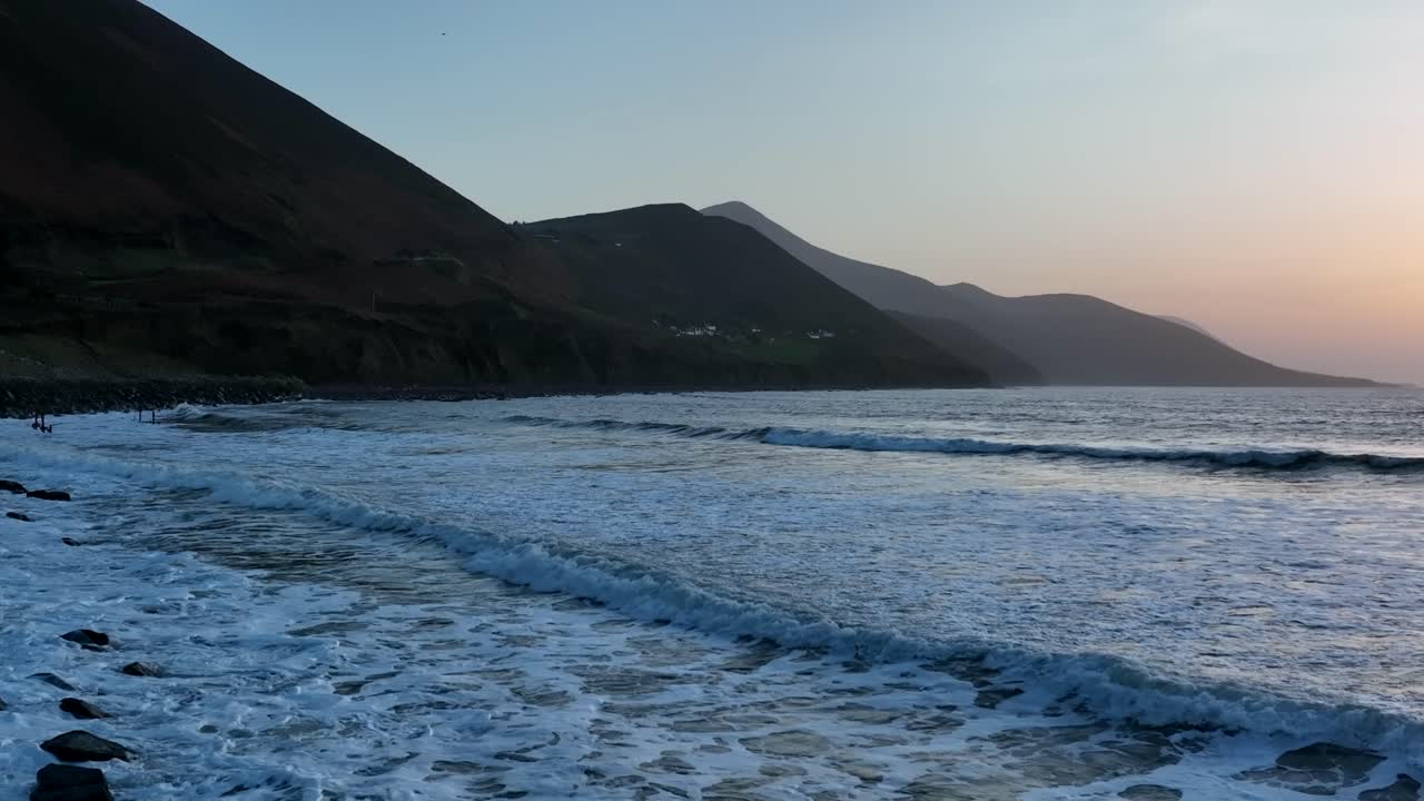 Spiaggia di Rossbeigh, Contea di Kerry, Irlanda, marzo 2022. Un drone si muove lentamente verso destra sopra onde che si infrangono e si ritirano con risacca sulle rocce, con sagome di montagne sullo sfondo