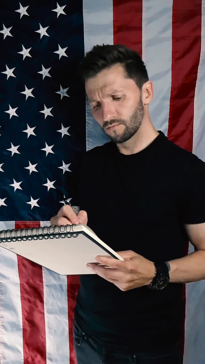 Tattooed white male writing down notes with a pen in a white notebook or notepad while listening to the viewer while standing and nodding in front of vertical United States of America flag. short hair