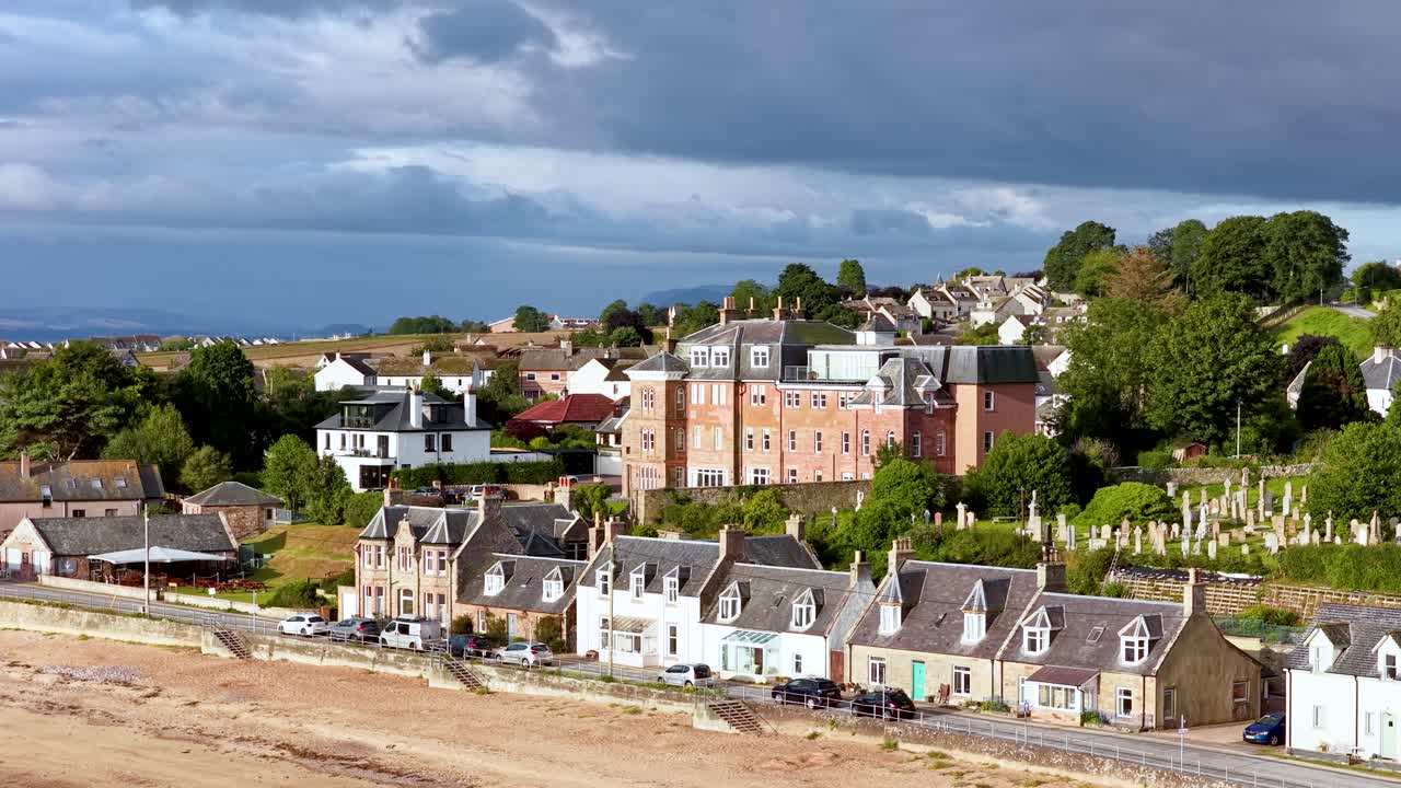 Drone pans across Rosemarkie village, beachfront houses, graveyard, and dramatic Highland landscape in sunlight