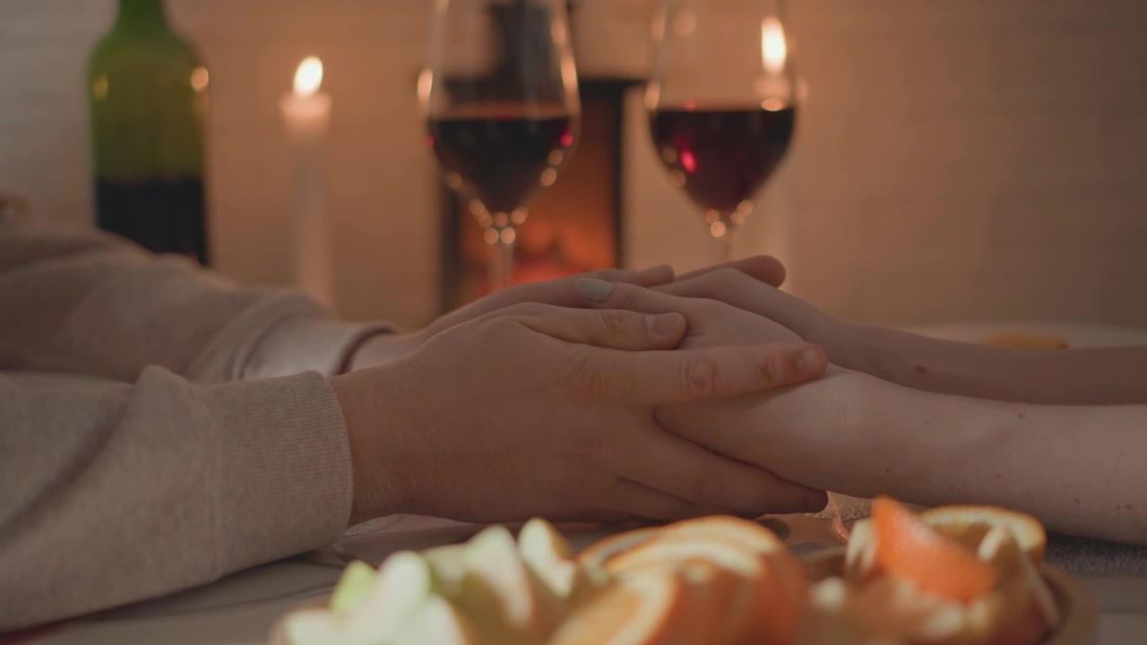 Vista ravvicinata di due persone che si tengono per mano sopra un tavolo da cena illuminato da candele, con vino, pasticcini, bicchieri e ombre calde che creano una connessione tenera, comfort e un'atmosfera serale romantica.