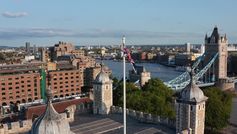 Union-Jack-slightly-waving-in-breeze.-Backwards-reveal-of-white-Tower-and-rest-of-Tower-of-London-castle-complex.-Tower-Bridge-across-River-Thames-in-background.-London,-UK