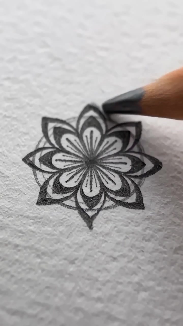 Una vista ravvicinata mostra una matita di grafite che crea un intricato disegno floreale all'interno di un contorno circolare, aggiungendo petali a strati e dettagli decorativi su una superficie bianca texturizzata con una morbida illuminazione naturale.
