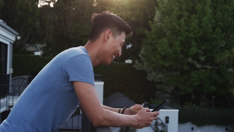 asian man smiling while using smartphone outdoors, enjoying sunny day, on balcony, copy space