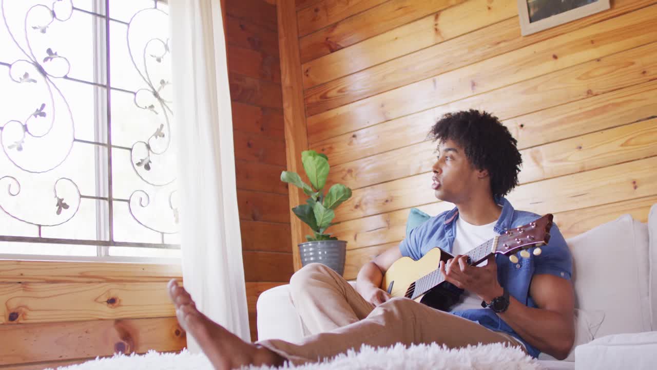 Uomo afroamericano felice seduto sul divano che suona la chitarra in una baita di legno, al rallentatore.