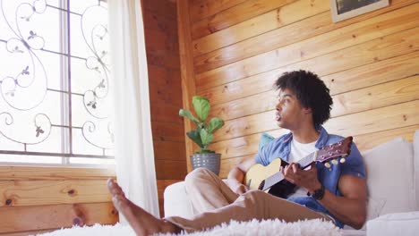 happy african american man sitting on sofa and playing guitar in log cabin, slow motion