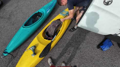 Overhead view of a man prepping kayaks beside a vehicle in bright sunlight. Scene captures the vibrant colors of the kayaks, set on the gravel, amid an adventurous atmosphere