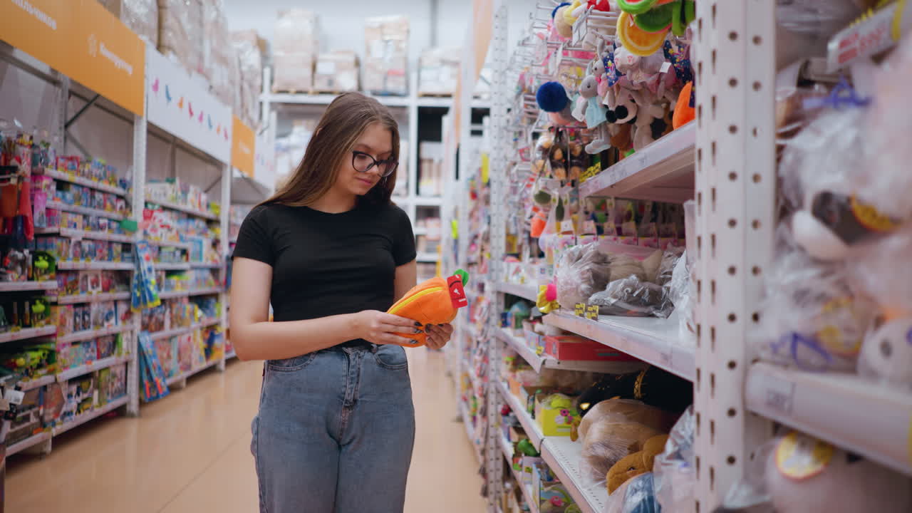 mujer joven en camiseta negra y vaqueros recogiendo una bolsa de peluche naranja brillante de un estante en una tienda de juguetes bien iluminada, juguetes coloridos y accesorios llenan los estantes mientras navega por las opciones