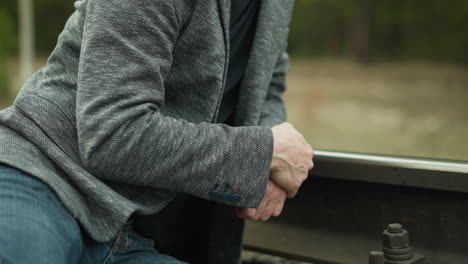 a close-up view of a man wearing a grey blazer and blue jeans, sitting and clasping his hands together on a railway track, as the camera moves away from him