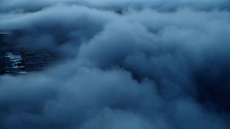 Thick cloud banks move over shadowed ridges and partially visible forested terrain, shifting from left to right as soft diffused light emphasizes rolling layers of fog and depth.
