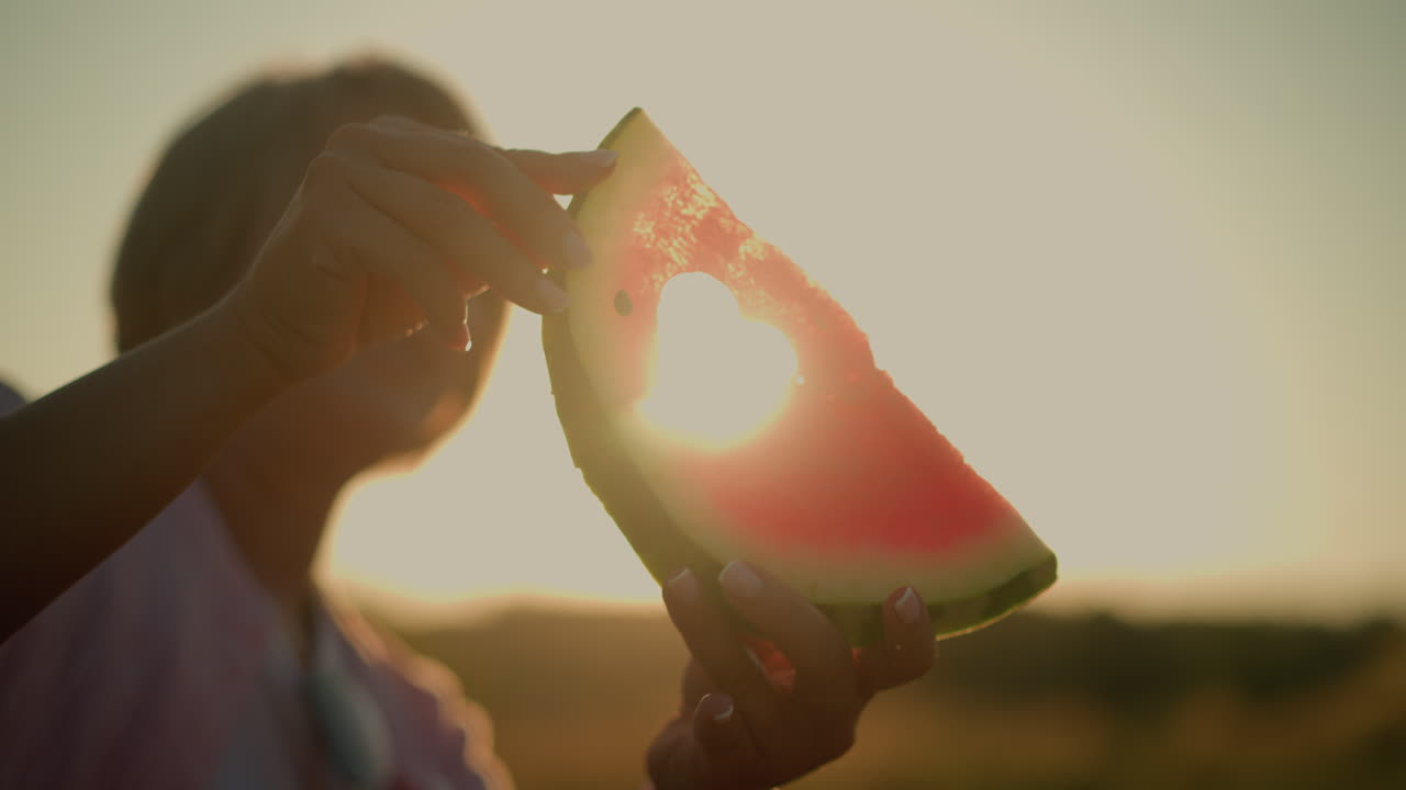 밝은 빛에 맞춰 심장 모양의 수박 조각을 들고 있는 여성의 클로즈업, 활기찬 은 과일을 조명하고 따뜻하고 만적인 실루을 만니다.