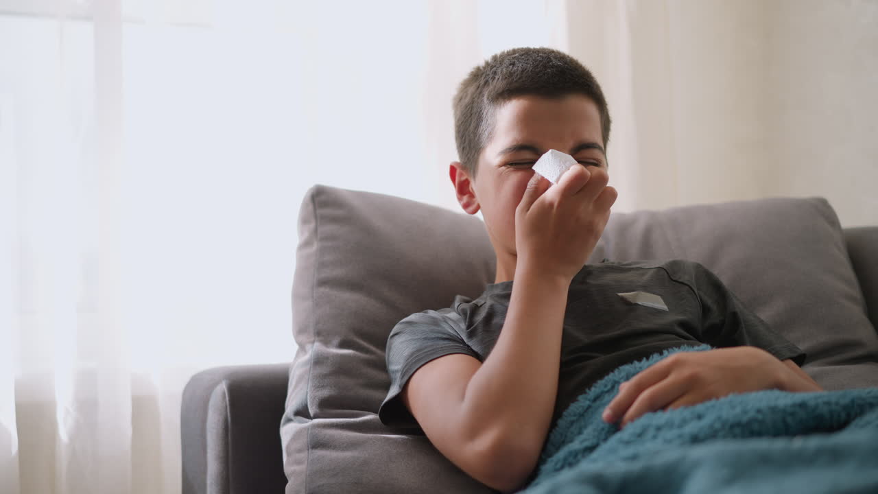 Sick student at home due to flu sneezing into tissue, resting on couch with blanket, light streaming through window, tissue littered around, glass cup on table