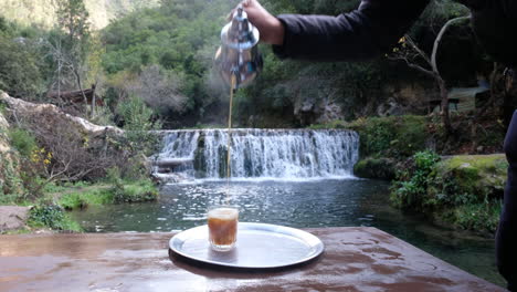 macho adulto sirviendo un vaso de té fresco frente a una cascada pacífica en marruecos