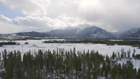 aerial moving forward to reveal icy, frozen lake surrounded by snow and green pine trees with large snowy ski town mountains in the background near silverthorne and frisco colorado