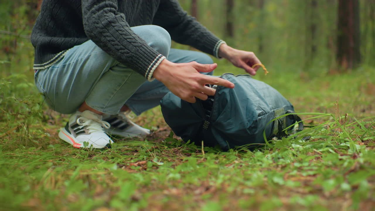 partial view of camper squatting on forest floor unzipping green outdoor bag while removing camping equipment with grass and dirt surrounding scene