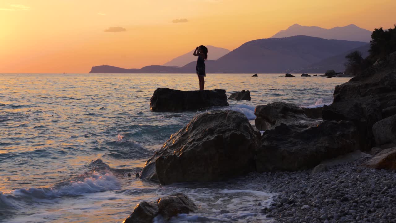 chica de pie en un acantilado bañado por las olas del mar en la hermosa puesta de sol con silueta de montañas y cielo naranja