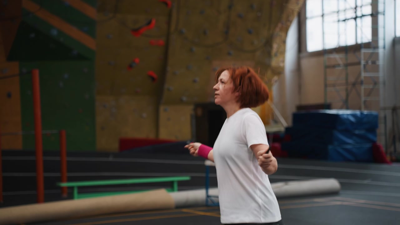 Indoor gym white redhead warming for badminton with skipping rope, steady rhythm and arm coordination, casual club practice vibe with mats and climbing wall, light conditioning and breathing focus