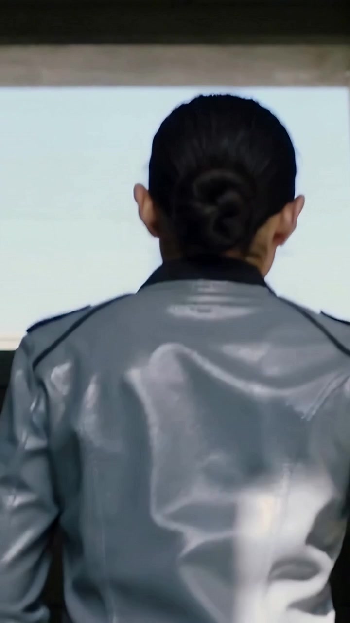 Vertical video: Walking woman in shiny jacket with bun, following bright light to doorway in hall