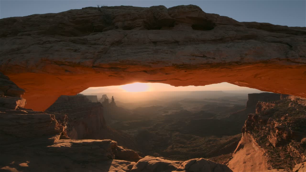 Solopgang gennem Mesa Arch, der skaber en fantastisk udsigt over ørkenlandskabet. Solens stråler skinner gennem klippeformationen og oplyser den enorme kløft og det klippefyldte terræn i det fjerne.