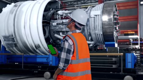 back view of asian male engineer with safety helmet working on a green screen smartphone and looking around while standing with airplane engine maintenance conducted, analytics checking the turbine