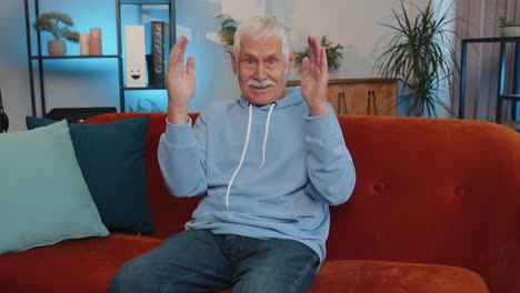 Senior-man-smiling-friendly-at-camera,-waving-hands-gesturing-hello,-hi,-greeting-at-home-on-couch