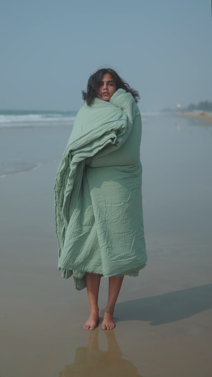 Full-body shot of woman wrapped in green blanket standing on windy beach